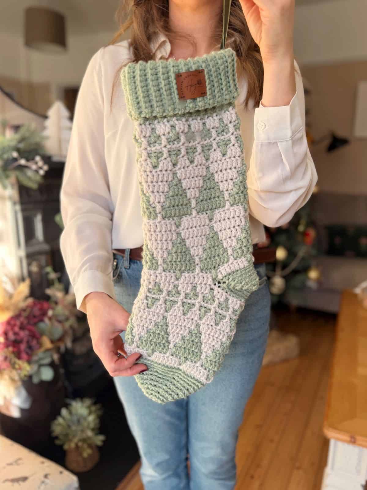 A person holds a large, crochet stocking patterned with green and white trees in a cozy living room.