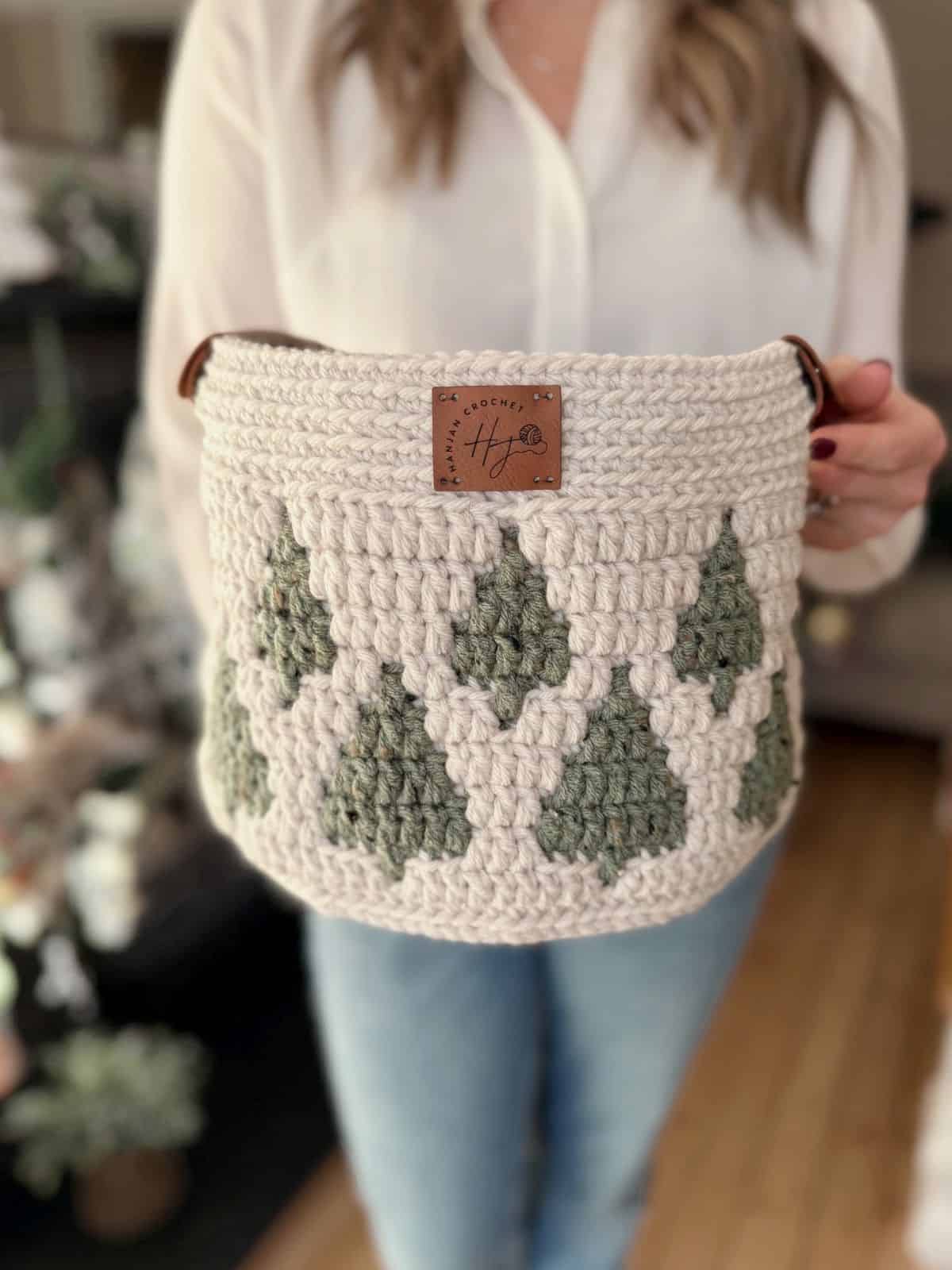 A person holds a christmas crochet basket with green and white geometric patterns and a brown label on the front.