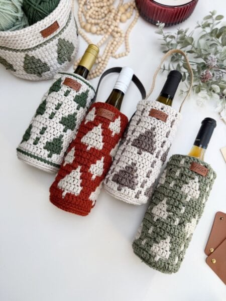 Four wine bottles in crochet wine bottle holders featuring tree patterns, displayed on a white surface with yarn, beads, and greenery in the background.