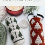 Two crochet wine bottle holders with tree patterns—one in green and white, the other in rust and white—are displayed alongside yarn, a candle, and a coaster.