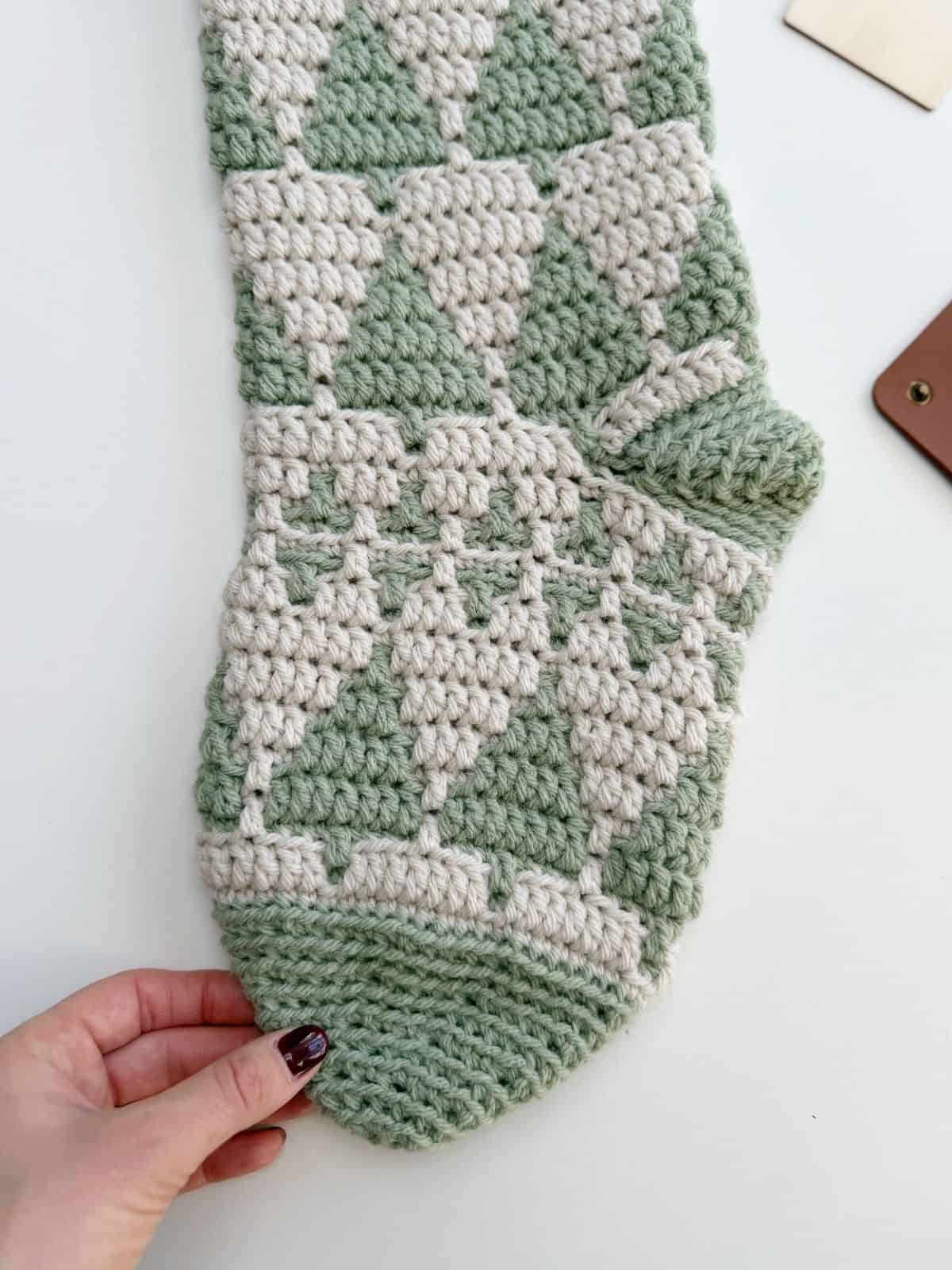 A hand holds the toe of a crocheted item with a green and white geometric pattern, lying on a white surface—perfect inspiration for your next crochet stocking pattern.