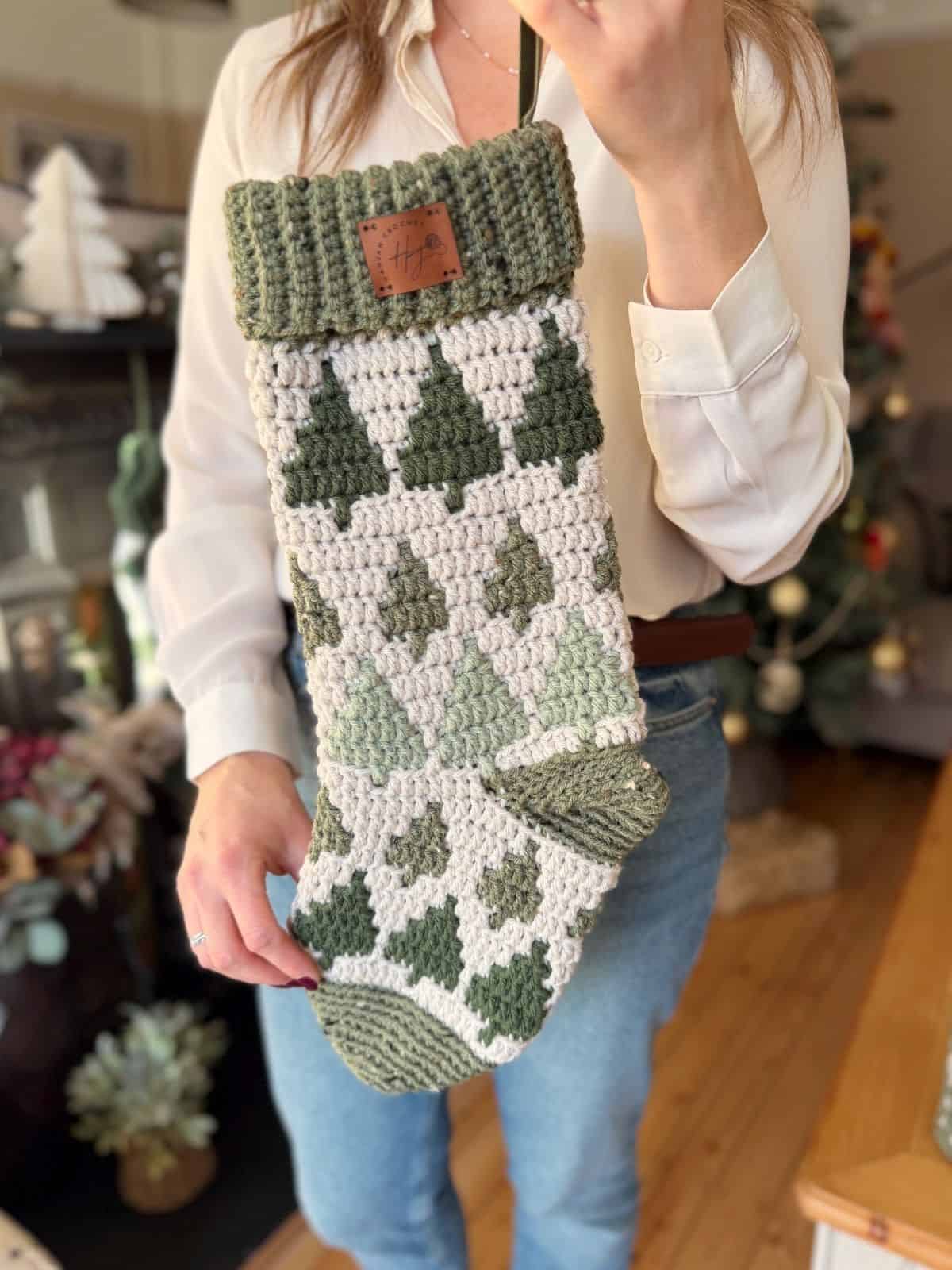 A person holds a large, crocheted Christmas stocking made with a festive crochet stocking pattern featuring green and white tree designs. A decorated Christmas tree and seasonal decor set a joyful background.