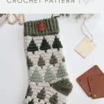 A crocheted Christmas stocking with a green tree pattern lies on a white surface next to blank tags and brown leather squares; text reads "crochet stocking pattern CROCHET PATTERN".