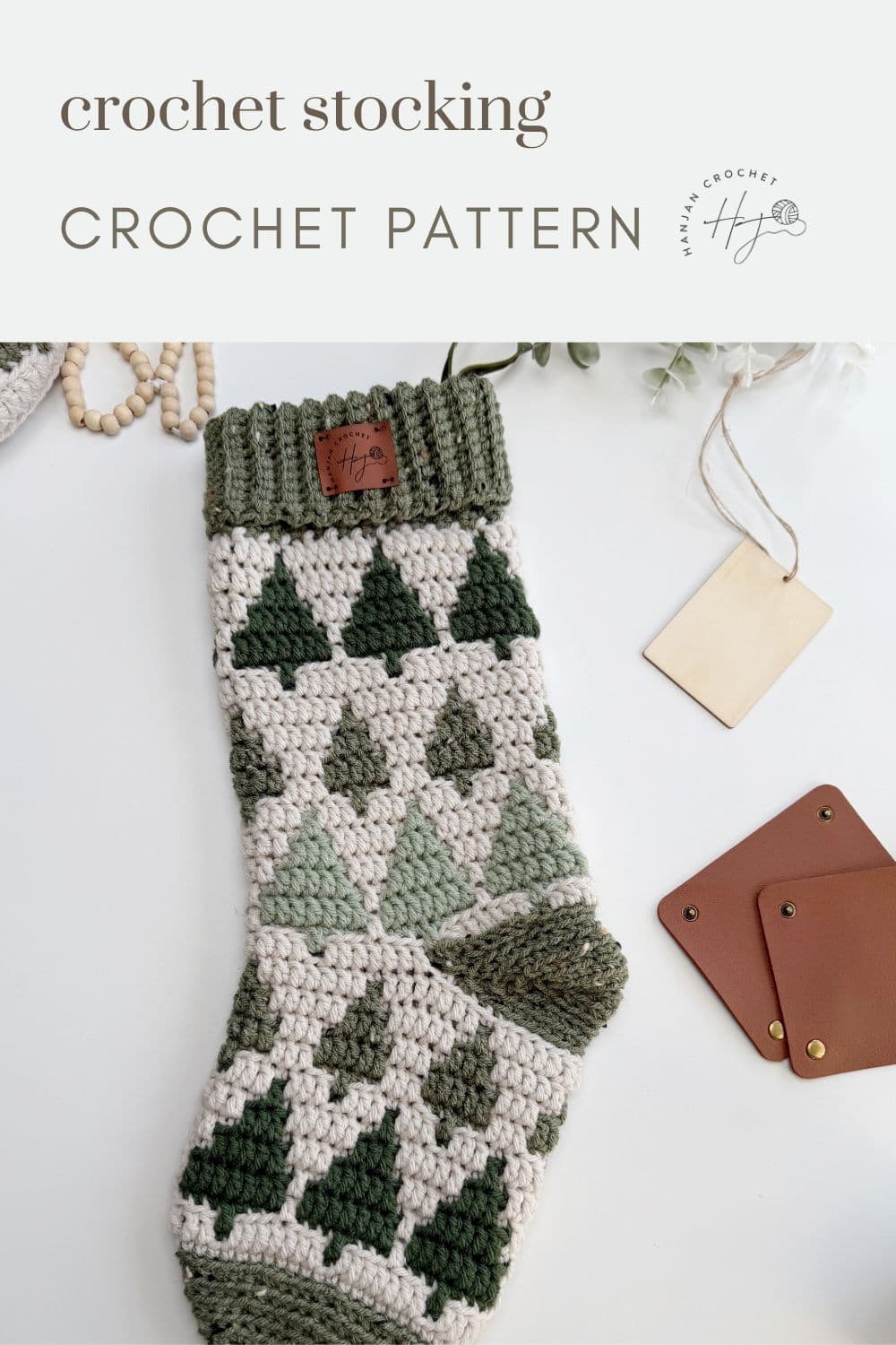 A crocheted Christmas stocking with a green tree pattern lies on a white surface next to blank tags and brown leather squares; text reads "crochet stocking pattern CROCHET PATTERN".