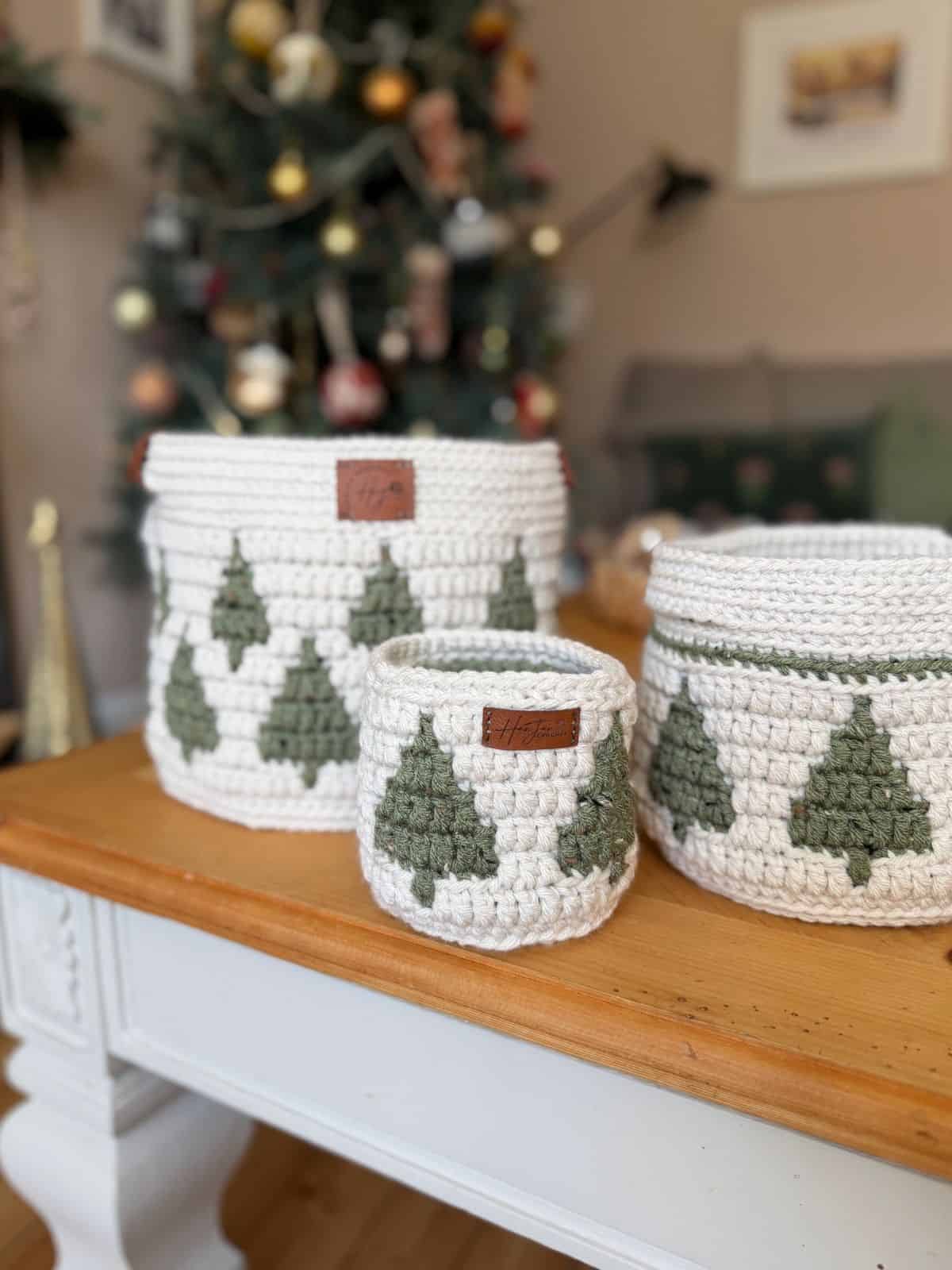 Three white christmas crochet baskets adorned with green Christmas tree patterns are displayed on a wooden table, with a decorated Christmas tree in the background.