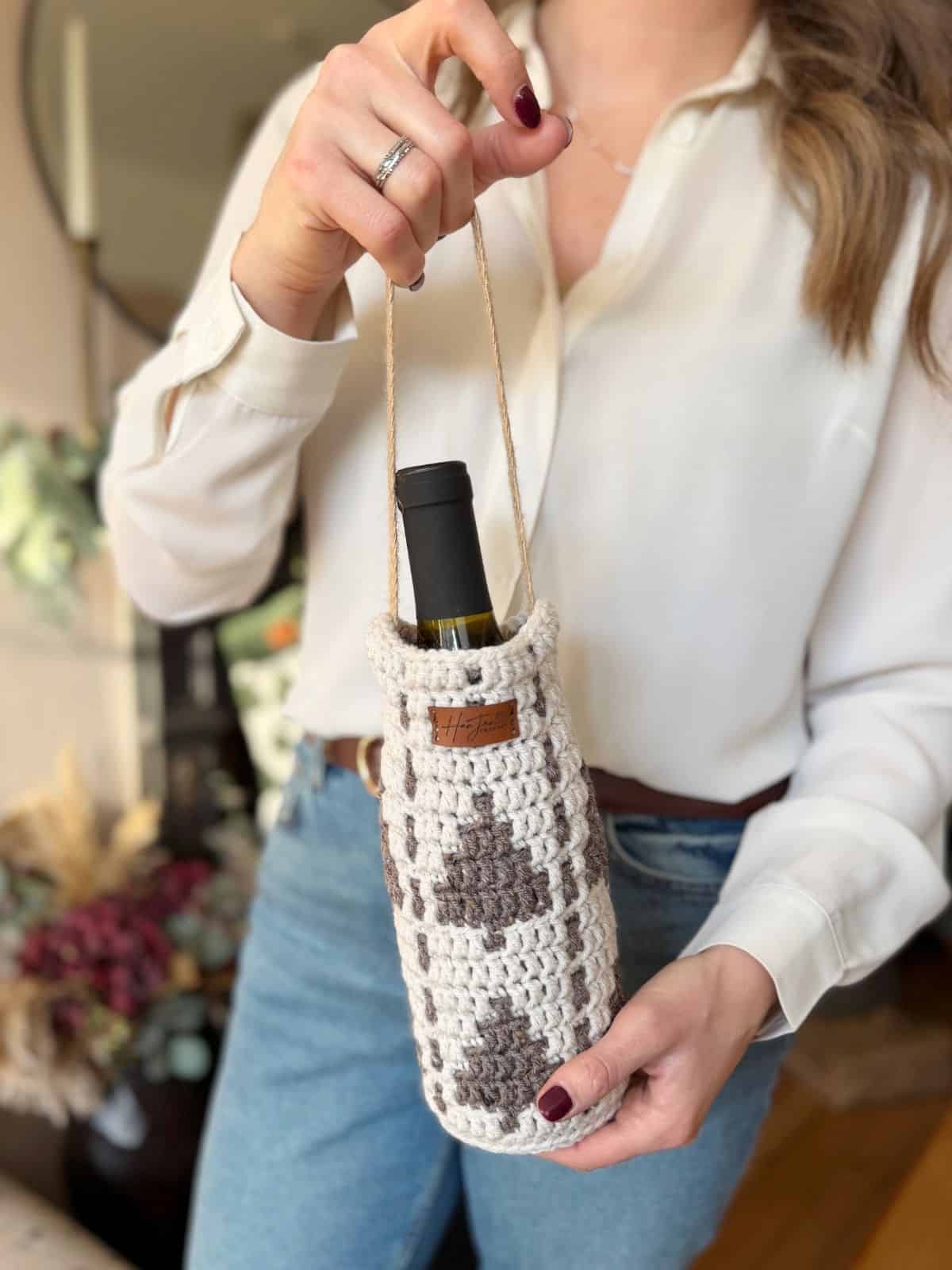 A person holds a wine bottle in a crochet wine bottle holder with a handle, dressed in a white shirt and blue jeans.