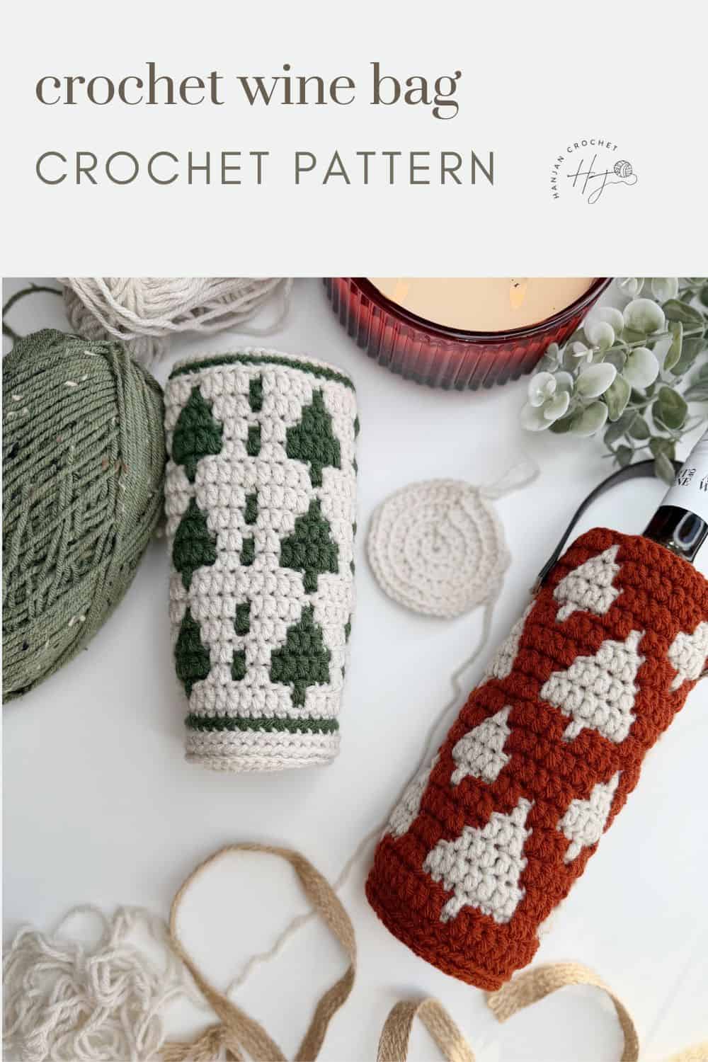 Two crochet wine bottle holders with tree patterns—one green and white, the other red and white—are displayed alongside yarn, a crochet hook, and a candle on a white surface.