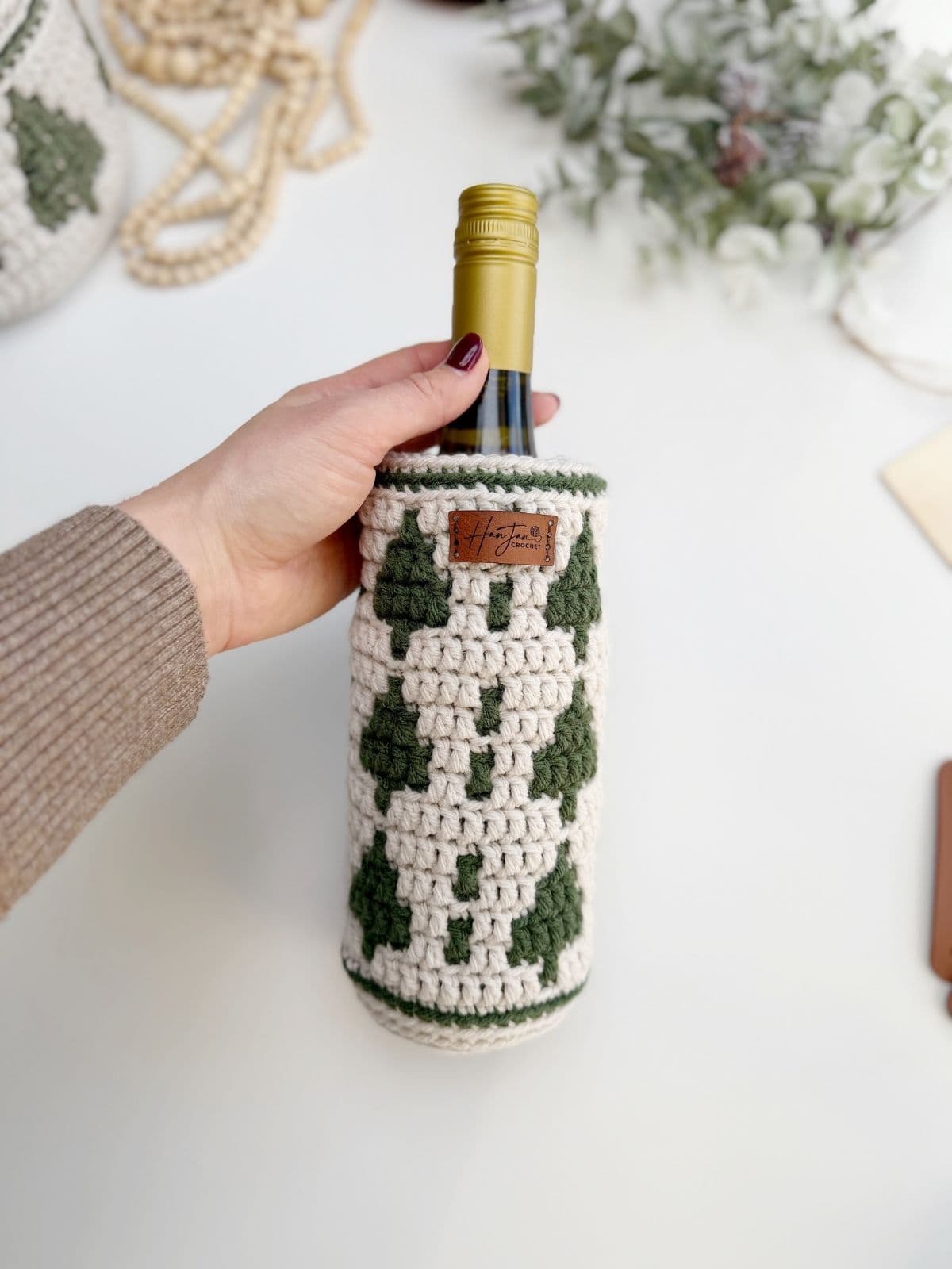 A hand holds a wine bottle wrapped in a crochet wine bottle holder, adorned with a green and white tree pattern.