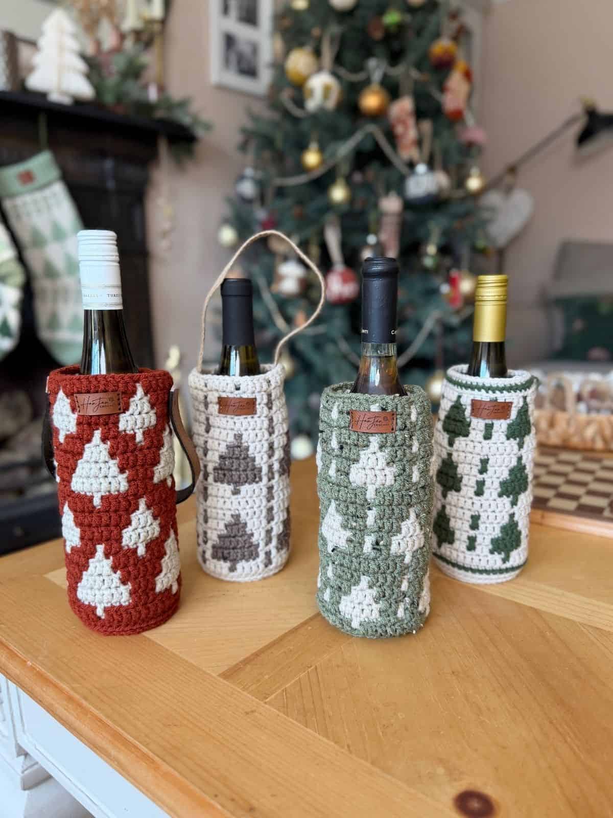 Four wine bottles in crochet wine bottle holders with tree patterns are placed on a wooden table in front of a decorated Christmas tree and festive home decor.