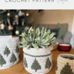 Three christmas crochet baskets with green tree patterns sit on a wooden table, one holding a potted plant. A decorated Christmas tree is visible in the background.