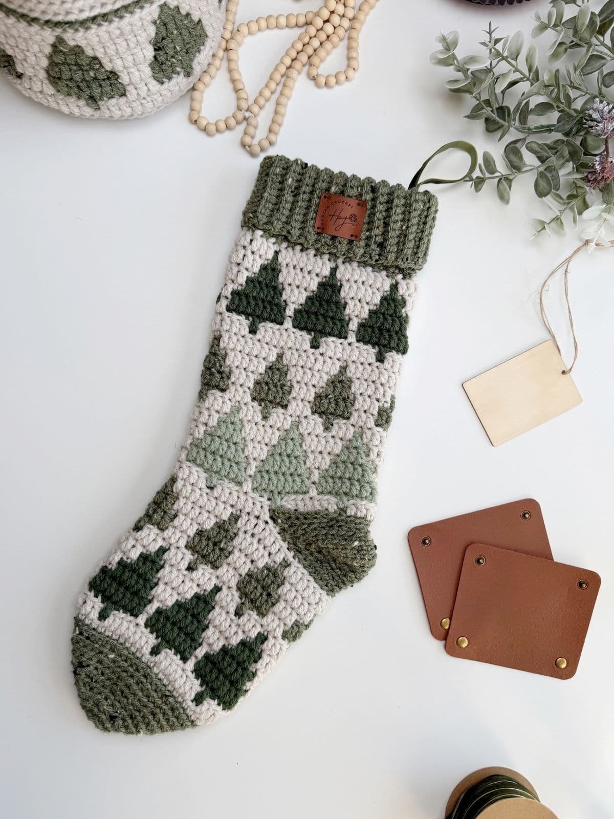 A crocheted Christmas stocking featuring a green and cream tree pattern lies on a white surface, surrounded by coasters, tags, beads, and greenery—perfect inspiration for your next crochet stocking pattern.