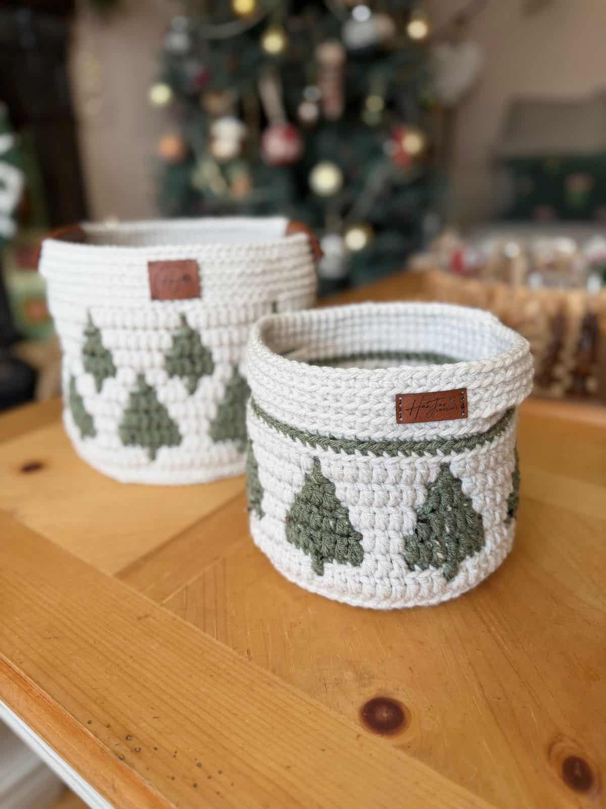 Two white christmas crochet baskets with green Christmas tree designs and brown leather tags sit on a wooden table; a decorated Christmas tree is visible in the background.