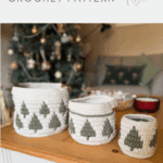 Three white christmas crochet baskets with green tree patterns are displayed on a wooden table in front of a decorated Christmas tree and a sofa.