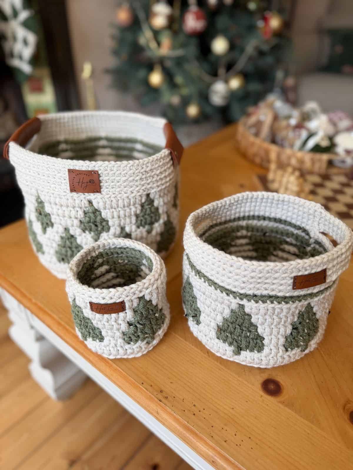 Three christmas crochet baskets with green tree patterns are arranged on a wooden table, with a decorated Christmas tree and another basket in the background.