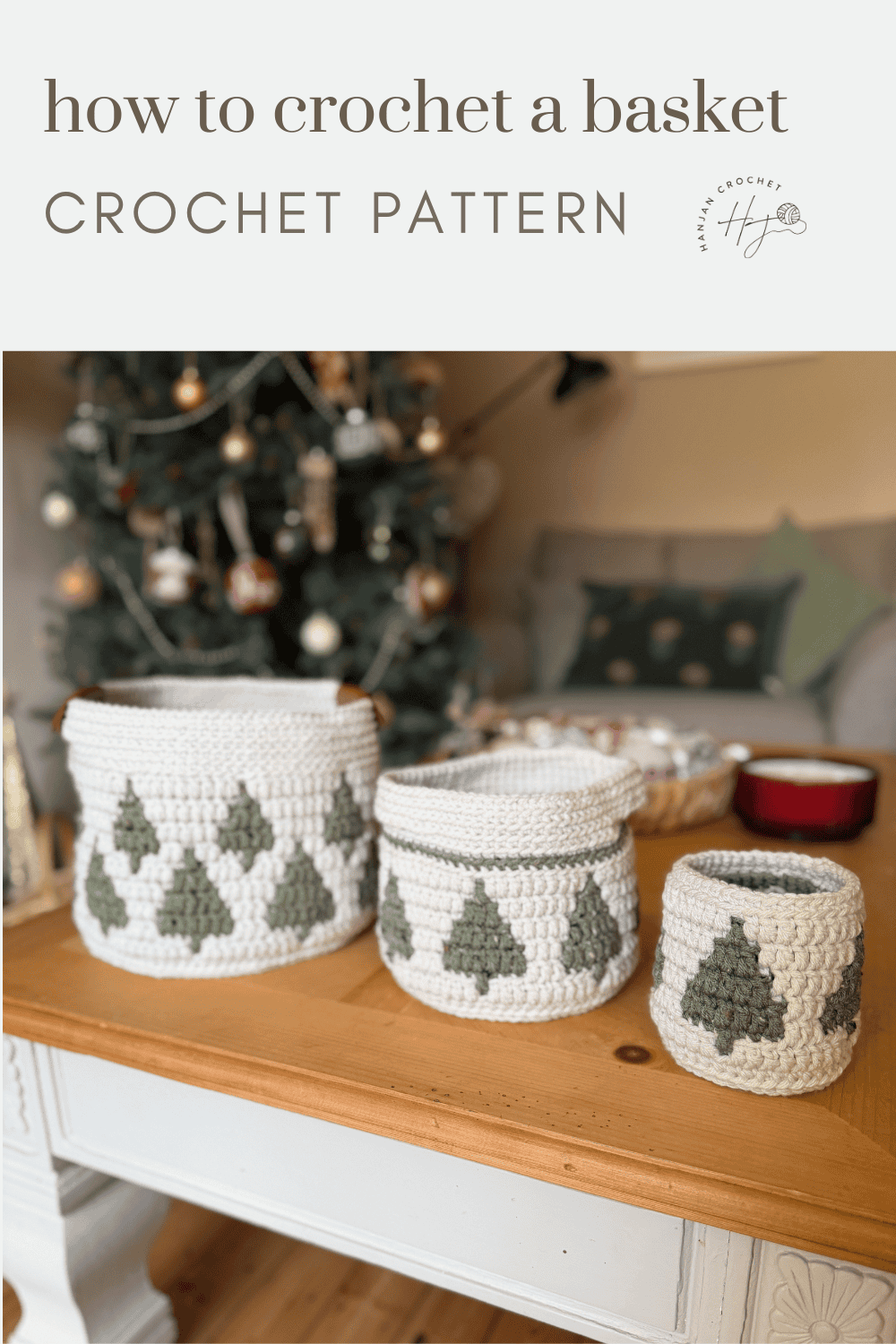 Three white christmas crochet baskets with green tree patterns are displayed on a wooden table in front of a decorated Christmas tree and a sofa.