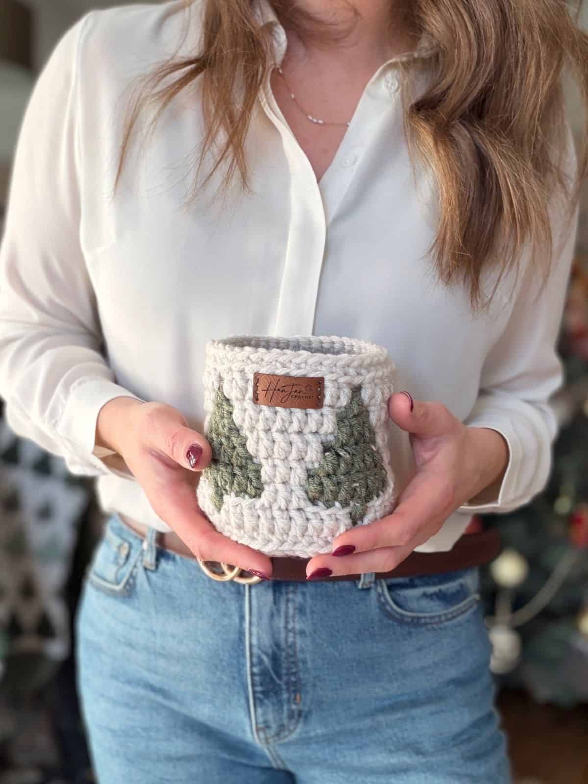 A woman in a white blouse and blue jeans holds a christmas crochet basket, featuring green and cream colors with a small brown label.