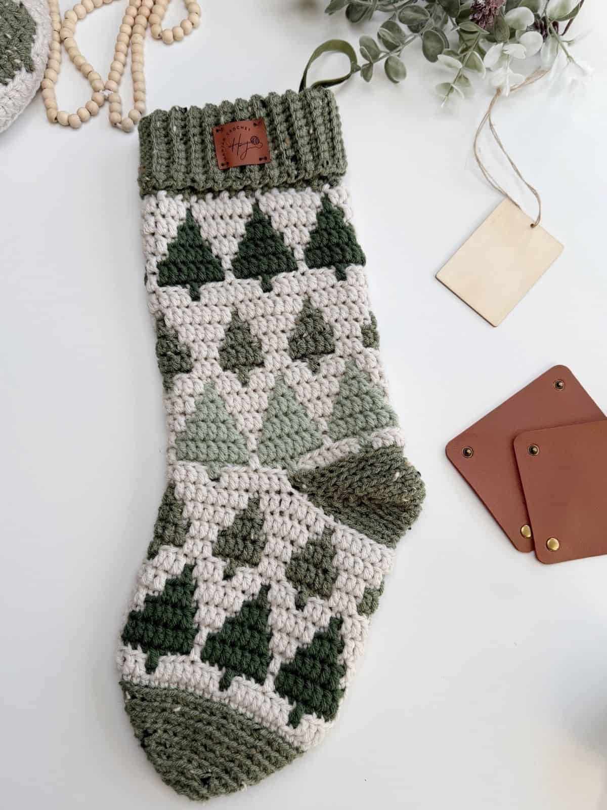 A crochet stocking pattern featuring green and white tree designs lies on a table near brown tags, a blank beige tag, and a beaded decoration with greenery.