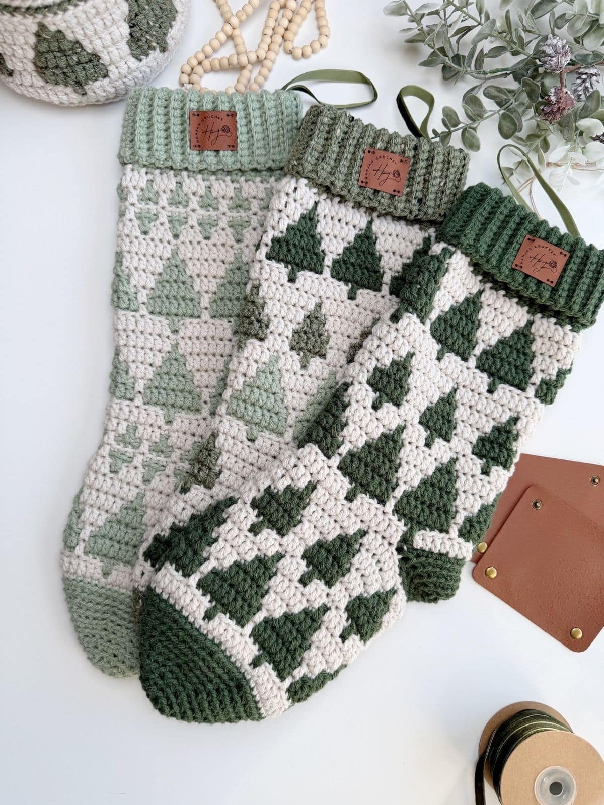 Three crocheted Christmas stockings with green and white tree patterns are arranged on a white surface, surrounded by craft supplies and greenery—a festive display perfect for showcasing your favorite crochet stocking pattern.