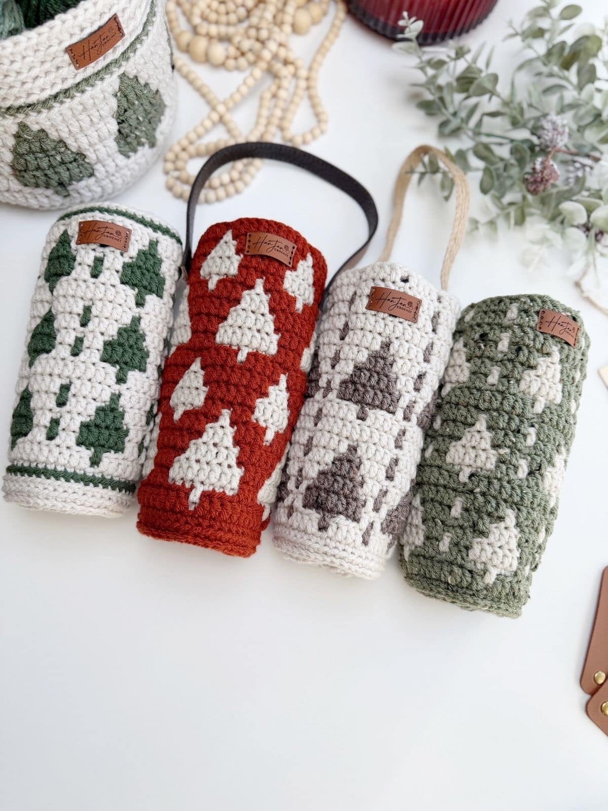 Four crocheted cylindrical pouches with tree patterns in green, red, grey, and sage—each doubling as a crochet wine bottle holder with a handy strap—are displayed on a white surface near plants and wooden beads.