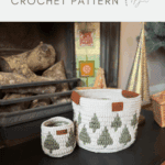 Two christmas crochet baskets with green Christmas tree patterns, one large with handles and one small, are displayed on a table in front of a cozy fireplace.