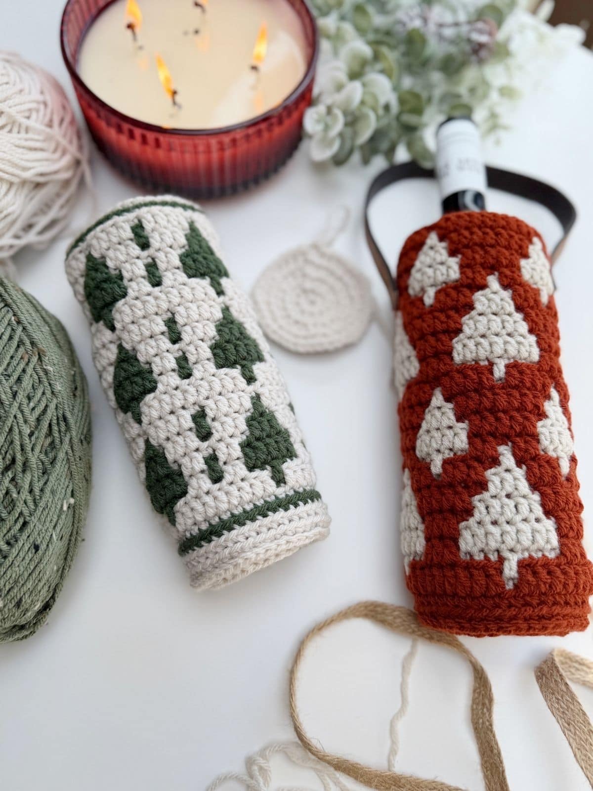 Two crochet wine bottle holders with tree patterns—one in green and white, the other in rust and white—are displayed among yarn balls, a candle, and crafting materials.