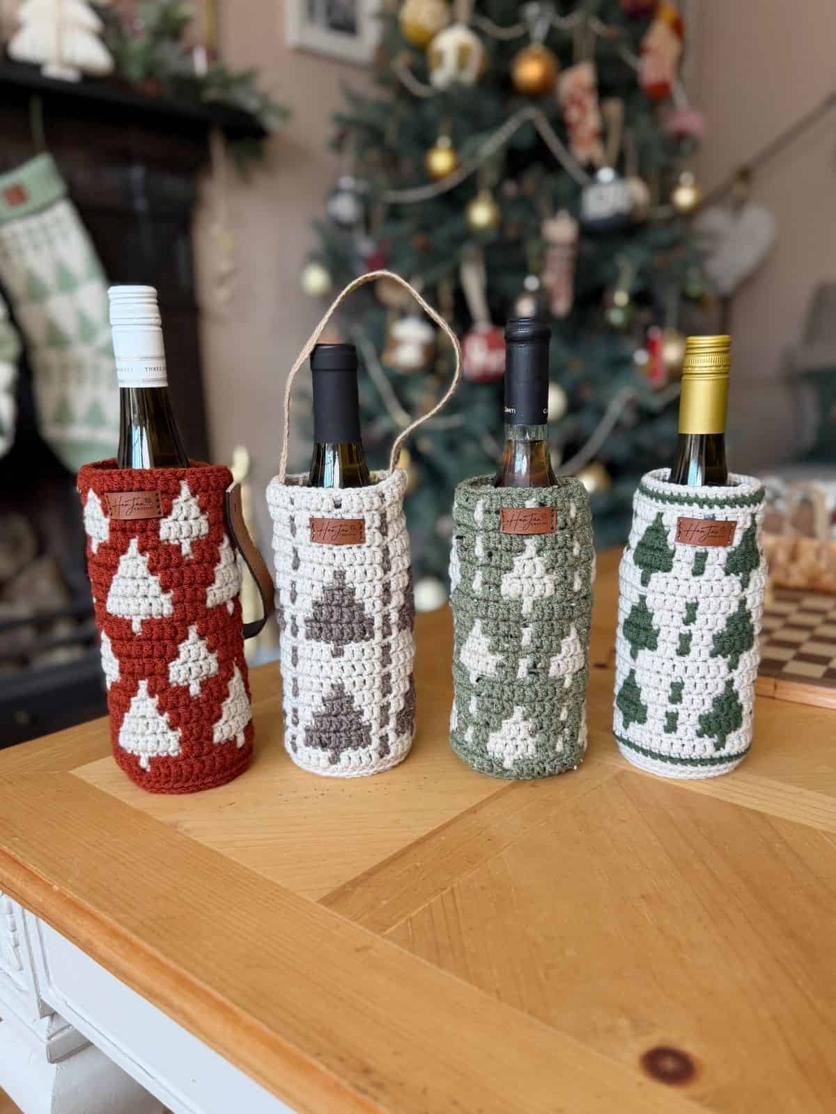 Four wine bottles in crochet wine bottle holders with tree patterns are arranged on a wooden table, with a decorated Christmas tree in the background.
