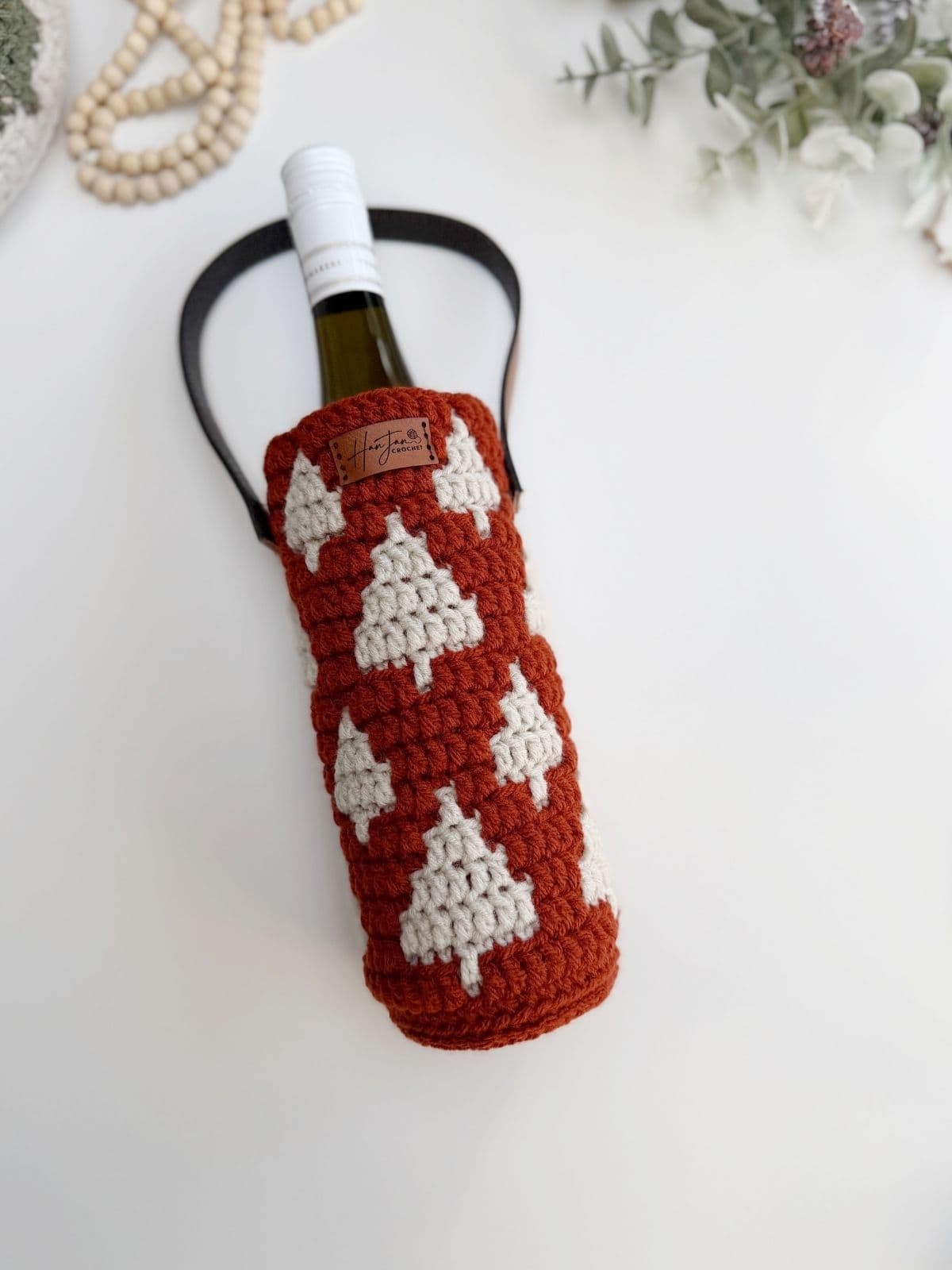 A crochet wine bottle holder in red with white tree patterns and a black handle, cradles a wine bottle on a white surface, accented by greenery in the background.