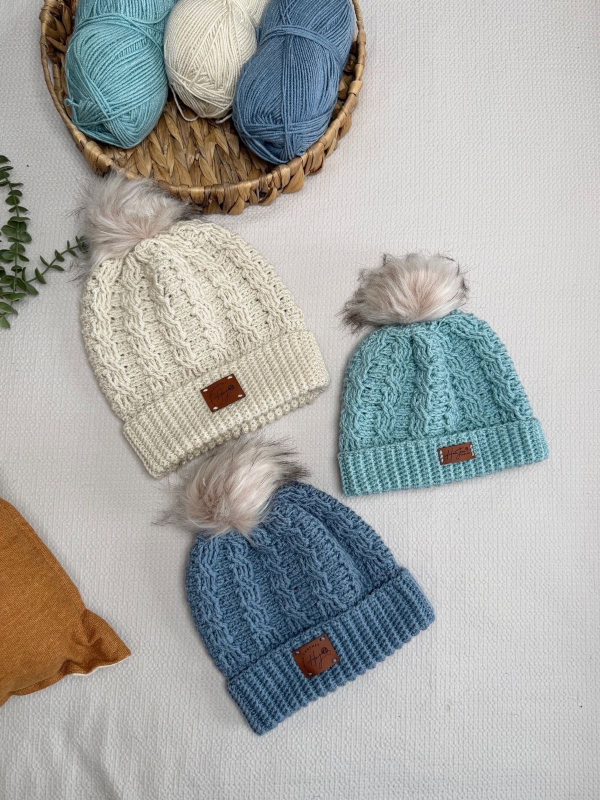 Three crocheted beanies with faux fur pom-poms—in cream, light blue, and blue—are arranged on a surface near a basket of yarn and a small green plant, perfect for inspiration or to pair with your favorite free crochet cable hat pattern.