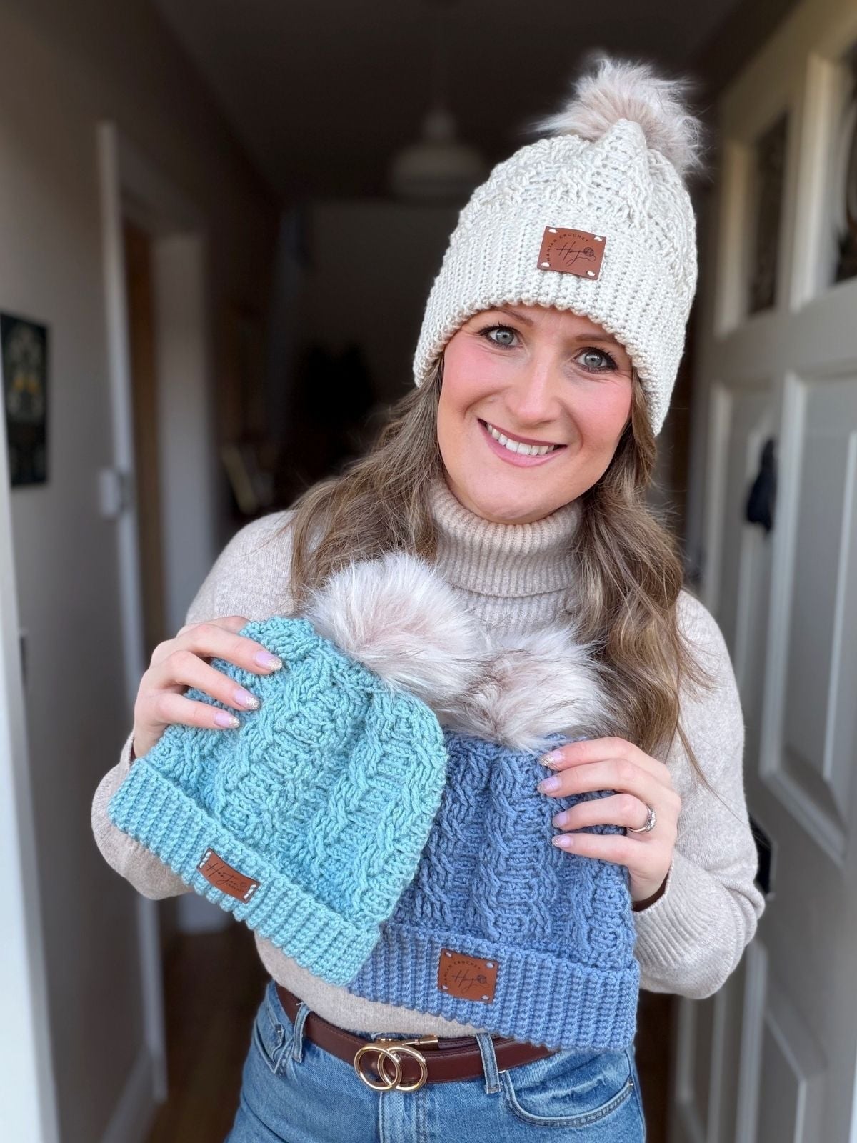 Woman in a cream crochet hat and beige sweater holds two blue crochet hats with faux fur pom-poms, showcasing her latest creations made from a free crochet cable hat pattern, standing indoors in a hallway.