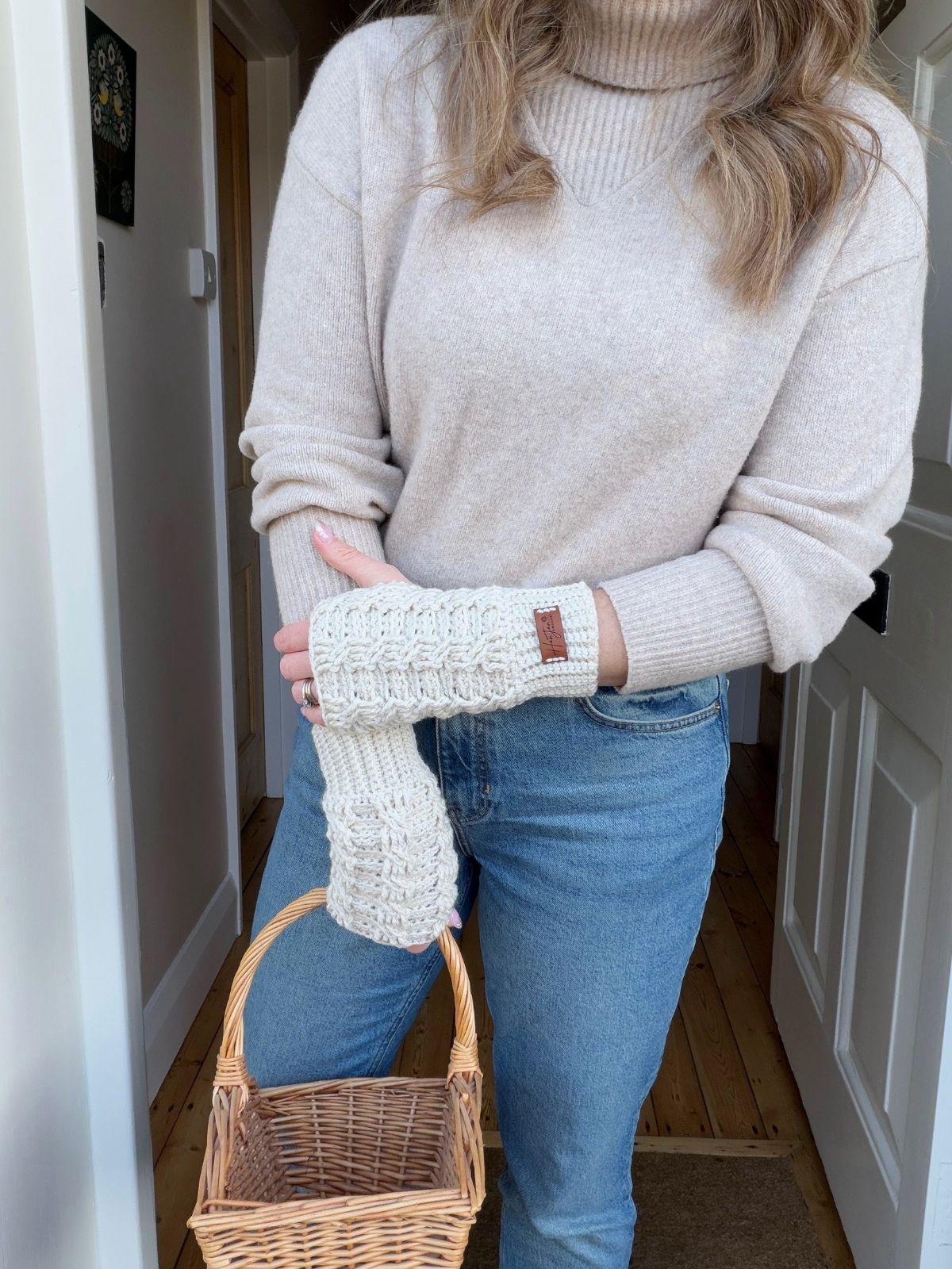 Woman wearing a beige turtleneck sweater, blue jeans, and crochet wrist warmers (find a free pattern online) holds a small empty wicker basket indoors.