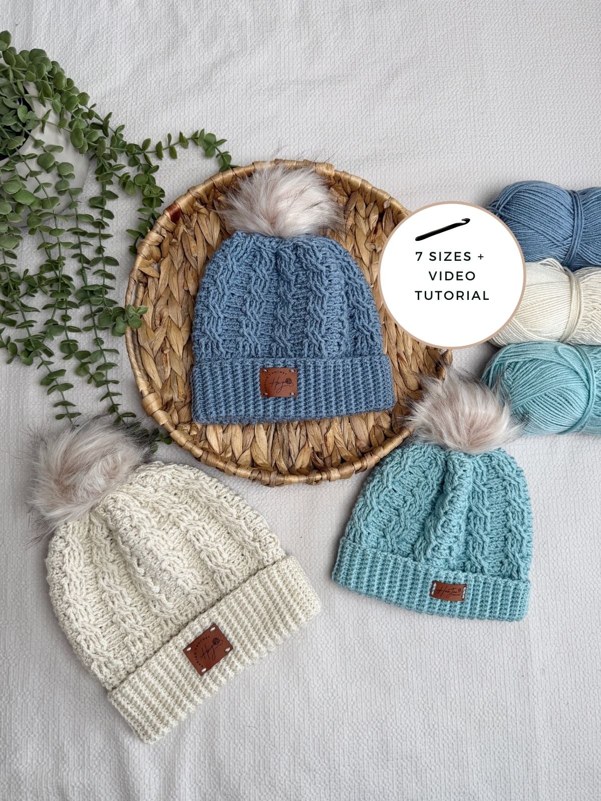 Three crochet hats with faux fur pom-poms are displayed on a woven mat and white surface, surrounded by yarn balls and greenery. A sign reads "7 sizes + video tutorial." Includes a free crochet cable hat pattern for added inspiration.