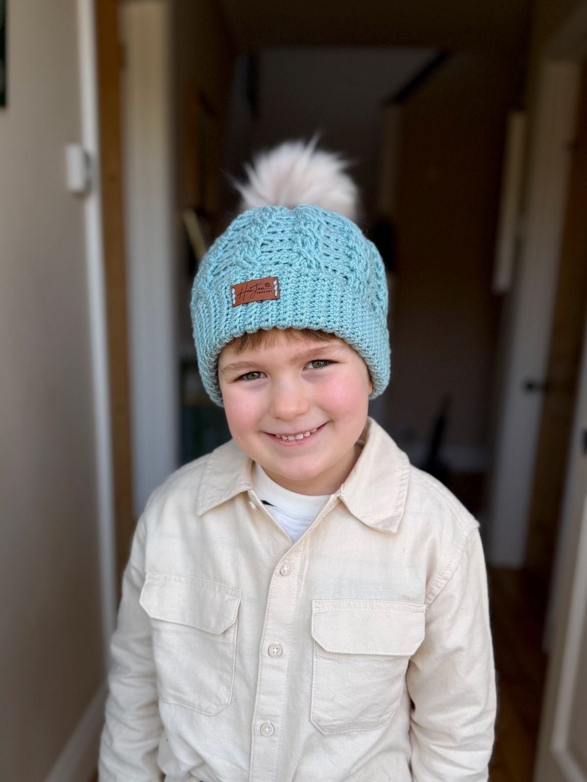 A young child wearing a light blue crochet beanie, inspired by a free crochet cable hat pattern, and a beige button-up shirt stands indoors, smiling at the camera.