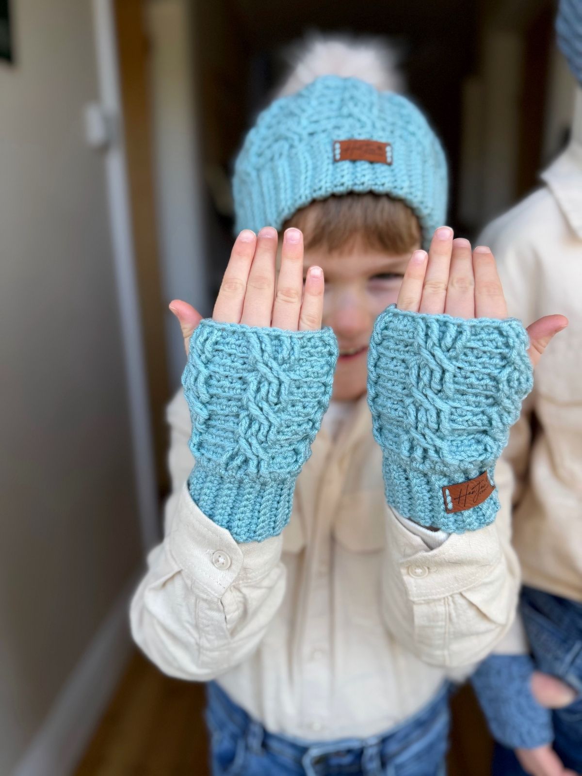 A child wearing a blue crochet hat and crochet wrist warmers from a free pattern holds both hands up, showing the gloves. The child is indoors and smiling.