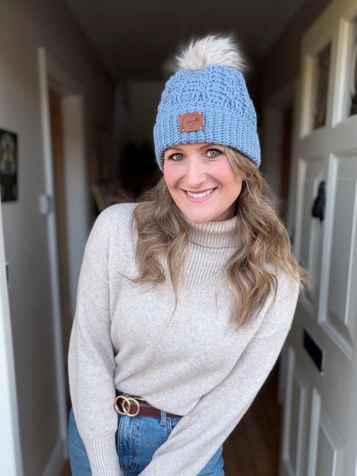 A woman wearing a light blue crochet hat with a pom-pom and a beige turtleneck sweater stands indoors, smiling at the camera, showcasing her cozy look inspired by a free crochet cable hat pattern.