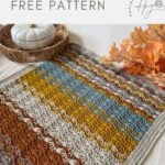 A multicolored lacy crochet blanket with mustard, blue, gray, and rust stripes is displayed on a table with a basket and autumn leaves. Text reads "lacy crochet blanket pattern free.