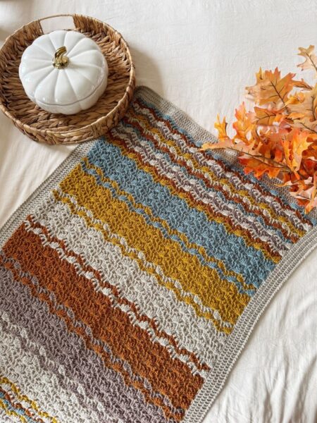 A crocheted blanket with horizontal stripes in autumn colors is laid on a cream surface next to a basket holding a white pumpkin dish and artificial orange leaves.