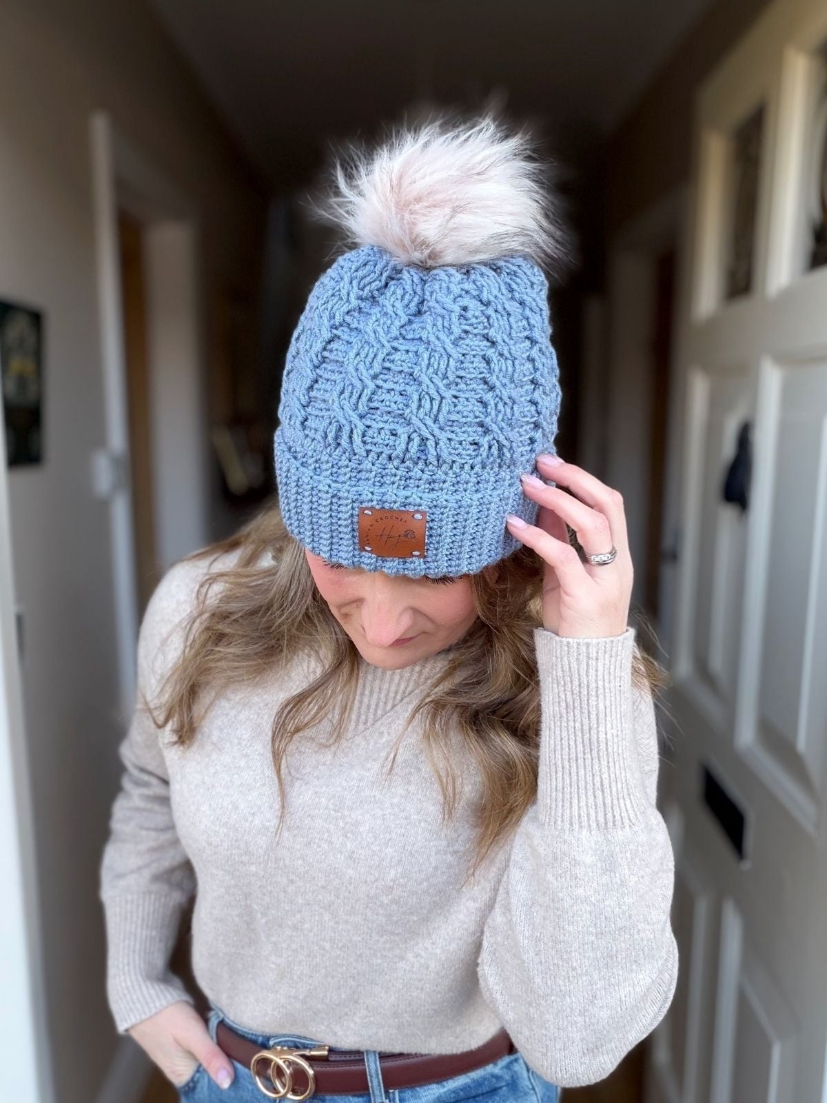 A person wearing a light blue crochet hat inspired by a free crochet cable hat pattern and a beige sweater stands indoors, looking down and touching the side of the hat.