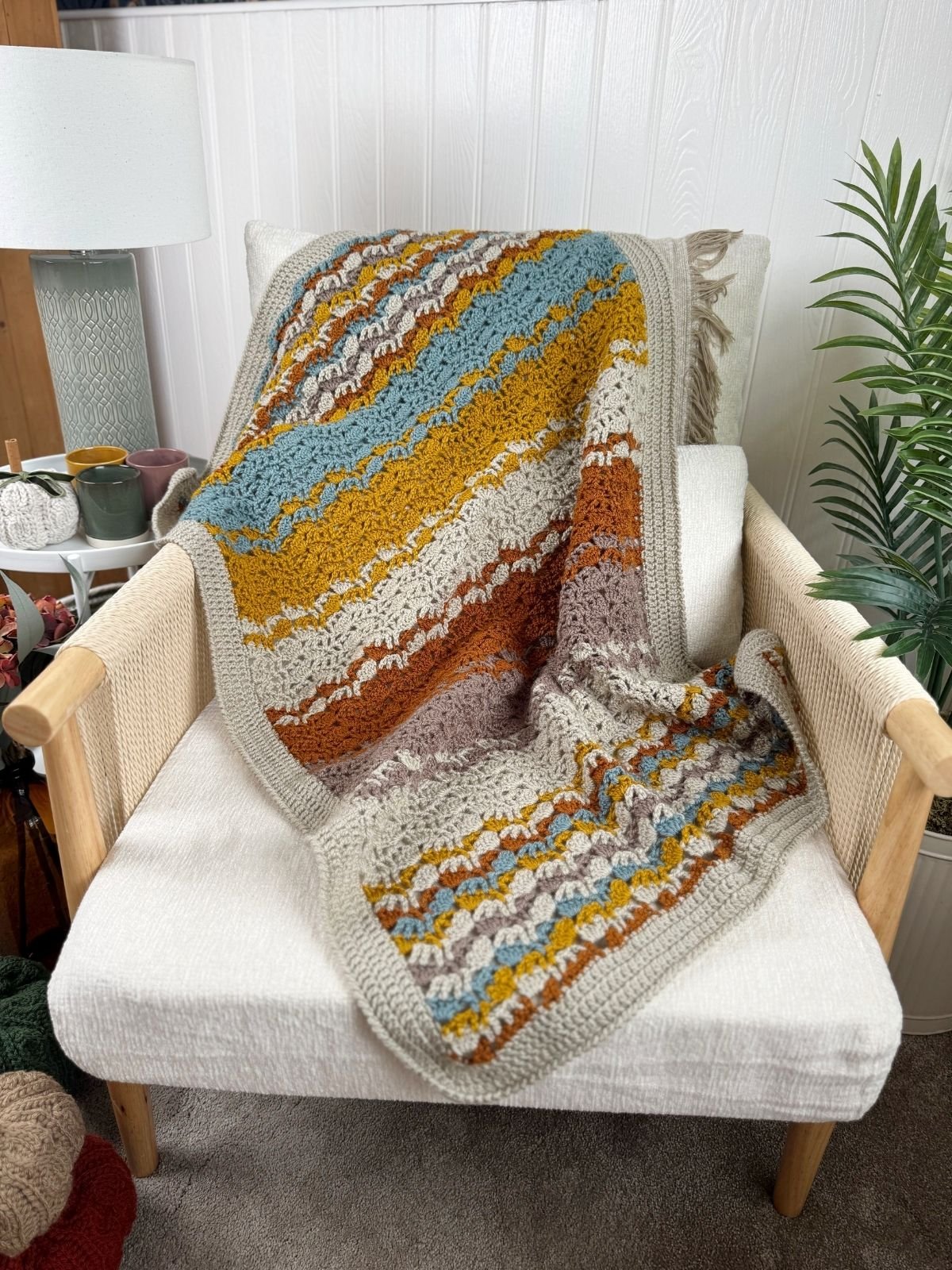 A multicolored lacy crochet blanket pattern with a striped design is draped over a cream armchair next to a potted plant and a side table with mugs and a lamp.