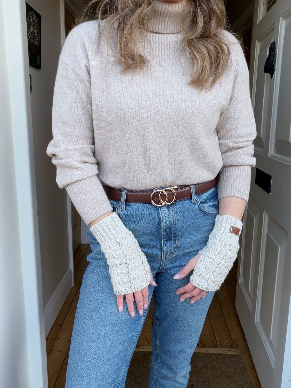 A person wearing a beige turtleneck sweater, blue jeans, a brown belt with a round buckle, and cream-colored crochet wrist warmers free pattern-inspired fingerless gloves stands indoors.