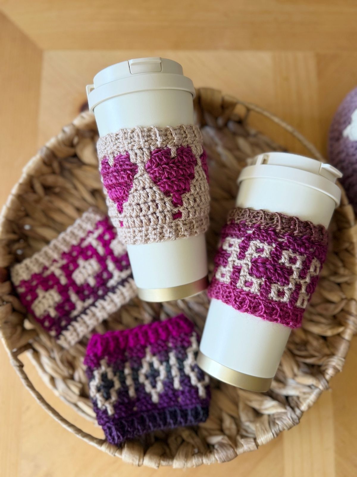 Two white travel mugs with crocheted cup sleeves in pink and purple patterns, including a crochet coffee cozy with heart, sit in a woven basket alongside two additional crochet sleeves.