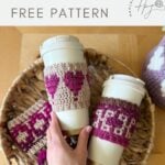 Two reusable coffee cups with crochet coffee cozies—one cozy adorned with a heart—are displayed in a woven basket alongside more handmade cozies. A hand is holding one of the cups.