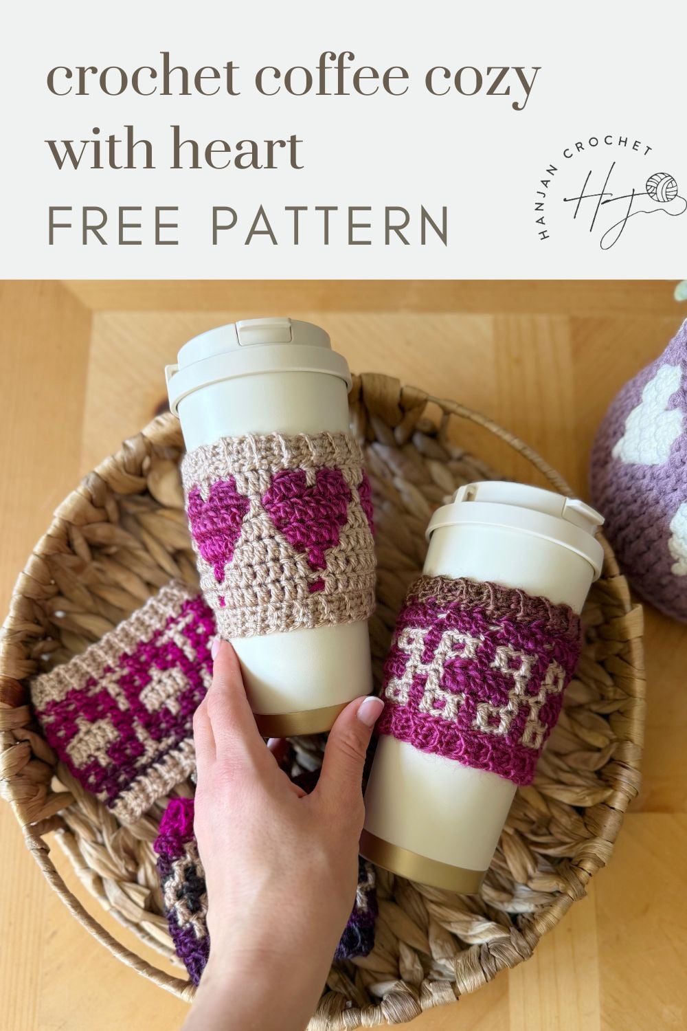 Two reusable coffee cups with crochet coffee cozies—one cozy adorned with a heart—are displayed in a woven basket alongside more handmade cozies. A hand is holding one of the cups.