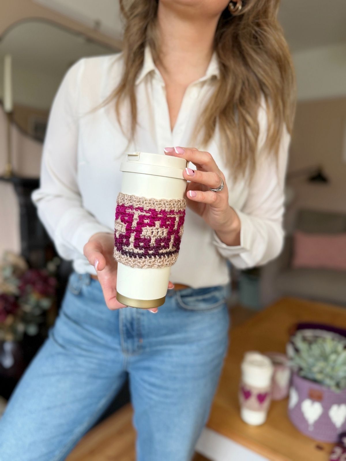 A person wearing a white shirt and jeans holds a reusable cup with a crochet coffee cozy with heart; another similar cup rests on a wooden table in the background.
