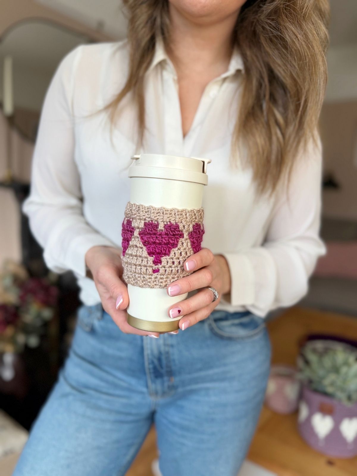 A person wearing a white blouse and blue jeans holds a reusable tumbler wrapped in a crochet coffee cozy with heart detailing.