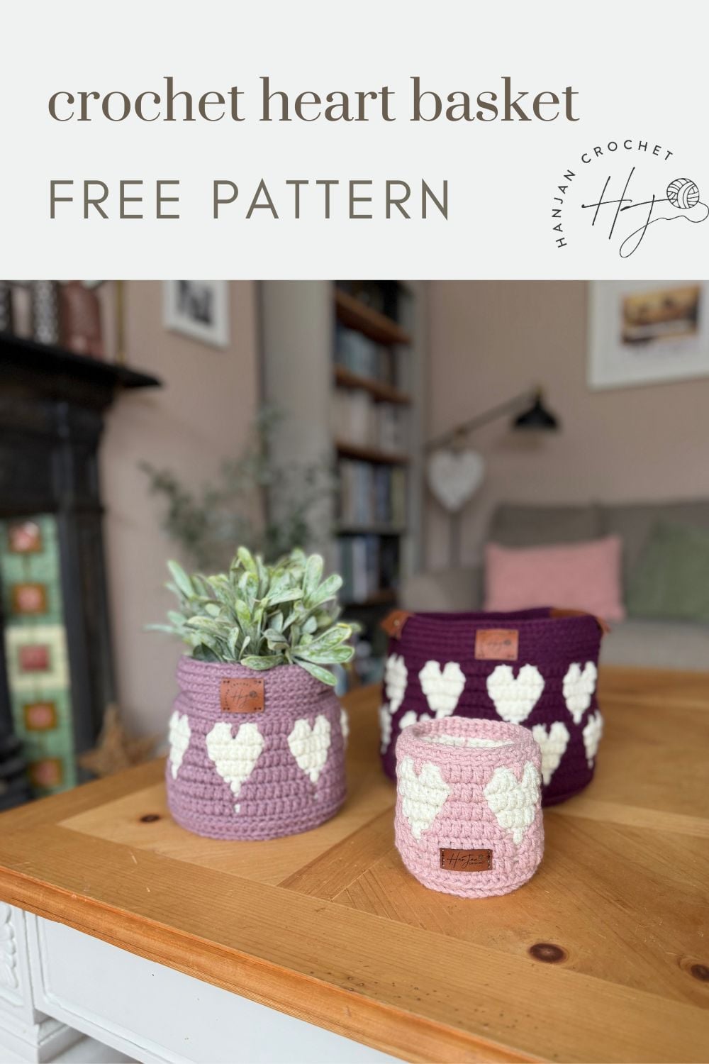 Three crochet baskets with white heart patterns, made using a crochet heart basket pattern, are displayed on a wooden table with a plant in one basket; a cozy living room is in the background.