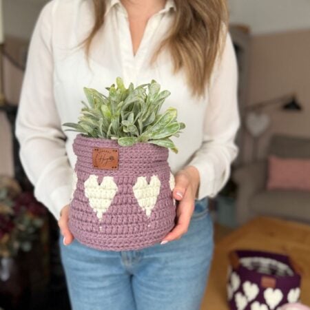 A woman in a white blouse and blue jeans holds a crocheted purple plant pot with white heart designs, featuring a charming crochet heart basket pattern, containing a green potted plant.