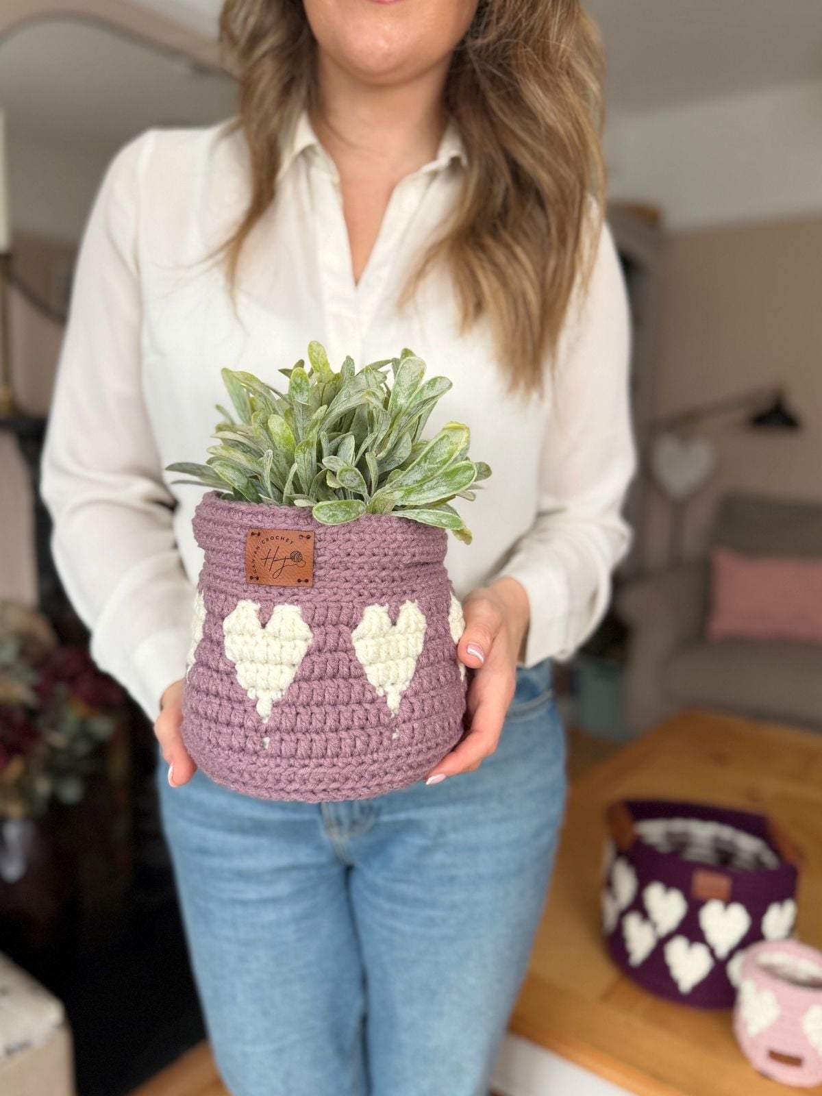 A woman in a white blouse and blue jeans holds a crocheted purple plant pot with white heart designs, featuring a charming crochet heart basket pattern, containing a green potted plant.