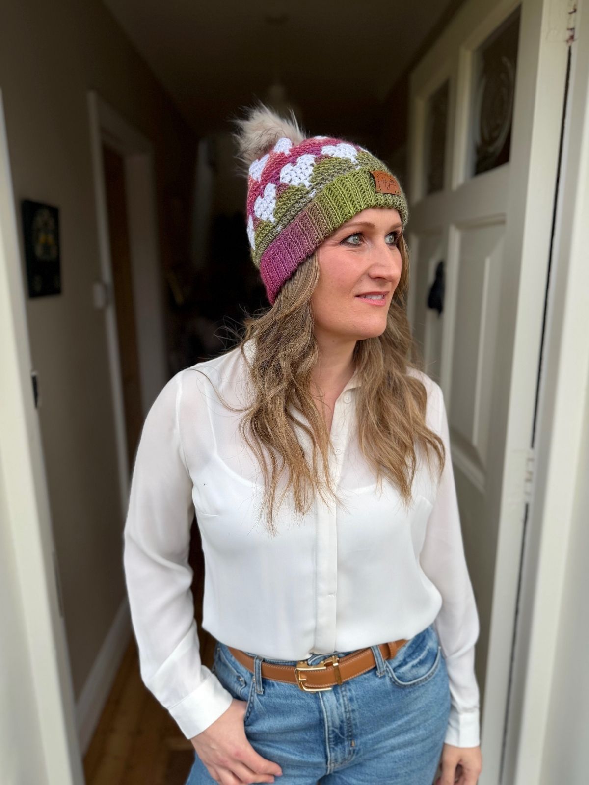 A woman in a white blouse, blue jeans, and a vibrant beanie made from a Free Crochet Heart Hat Pattern stands in a doorway, looking to the side.