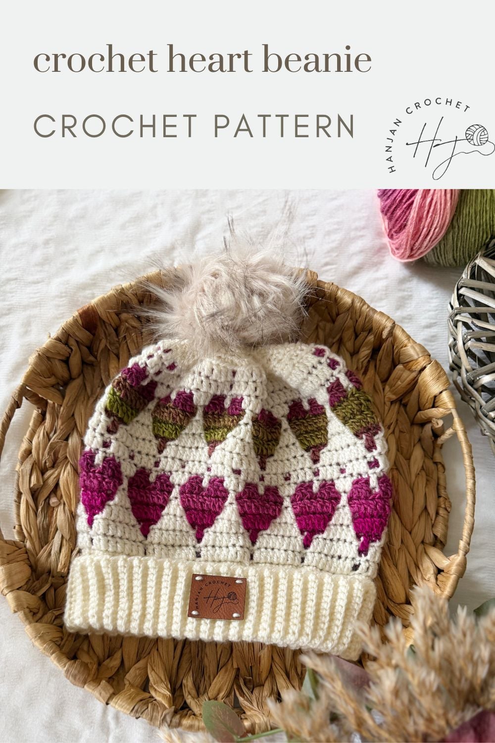 A crocheted beanie with pink and green heart patterns, topped with a faux fur pom-pom, displayed in a woven basket with yarn in the background. Text reads "Free Crochet Heart Hat Pattern CROCHET PATTERN.