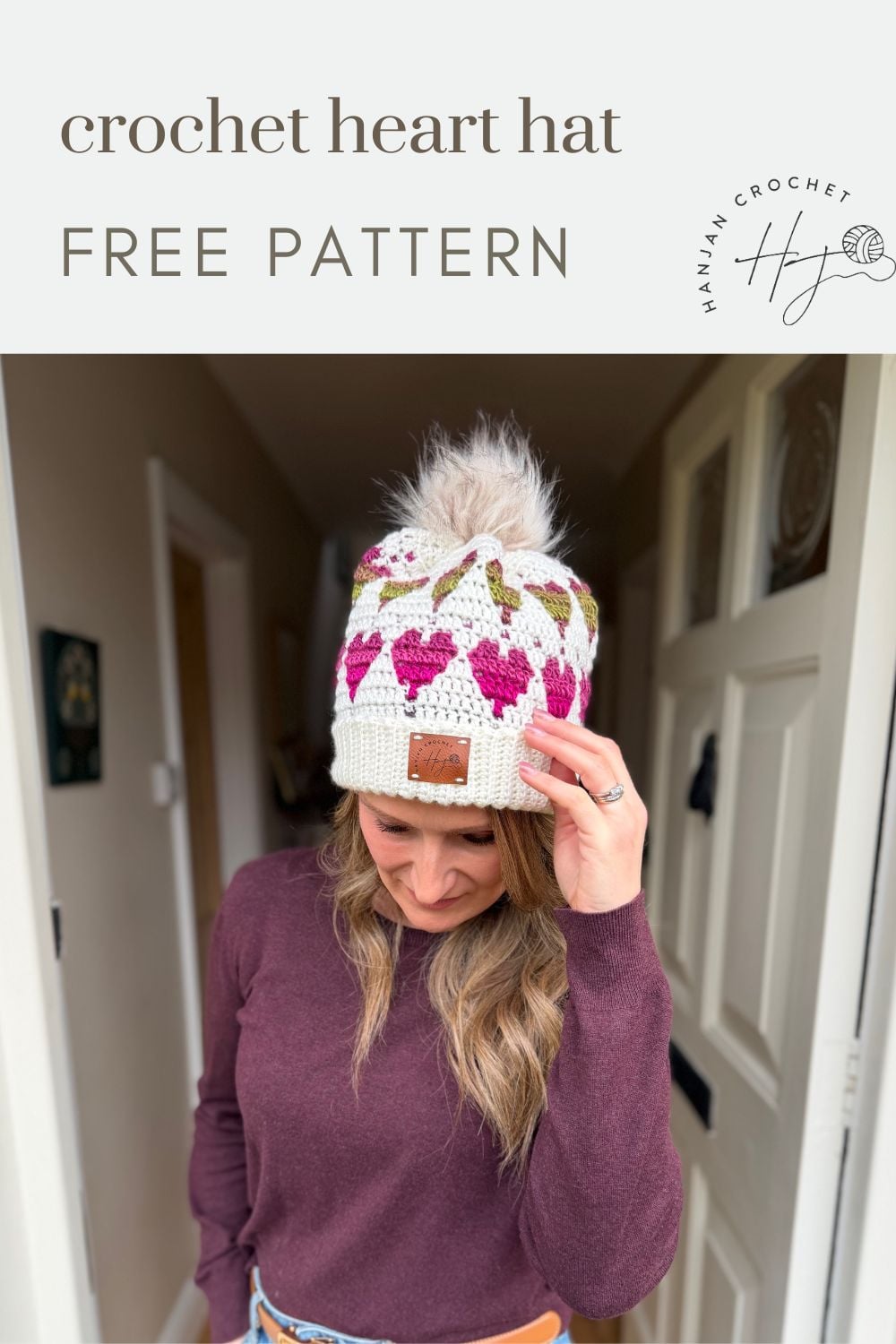 Woman wearing a white crochet hat with pink and green heart patterns and a pom-pom stands indoors under a sign that says "Free Crochet Heart Hat Pattern.